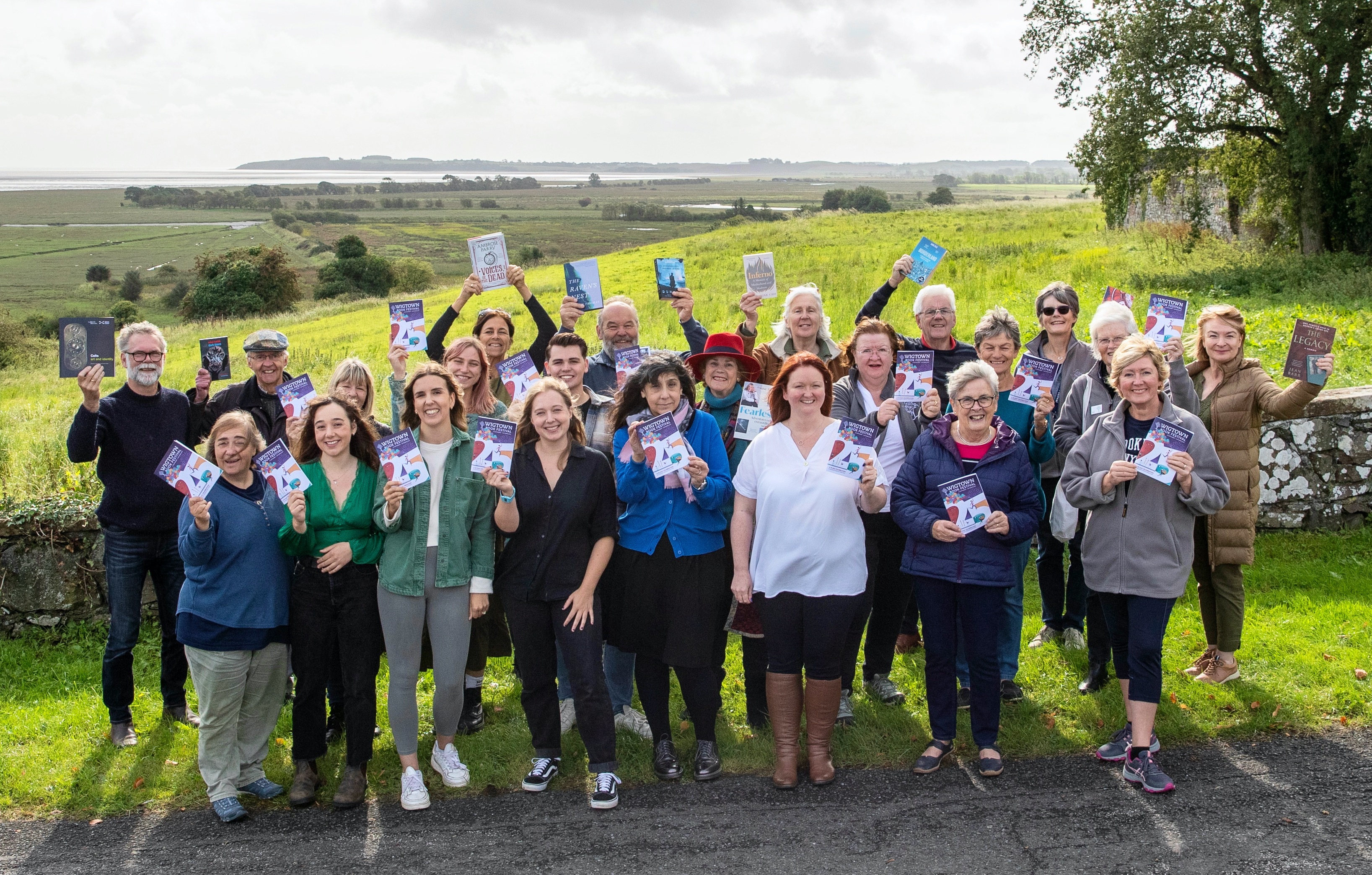 A crowd of staff and volunteers stand in front of a view over the Wigtown marshes. Each of them is holding a 2023 programme brochure.