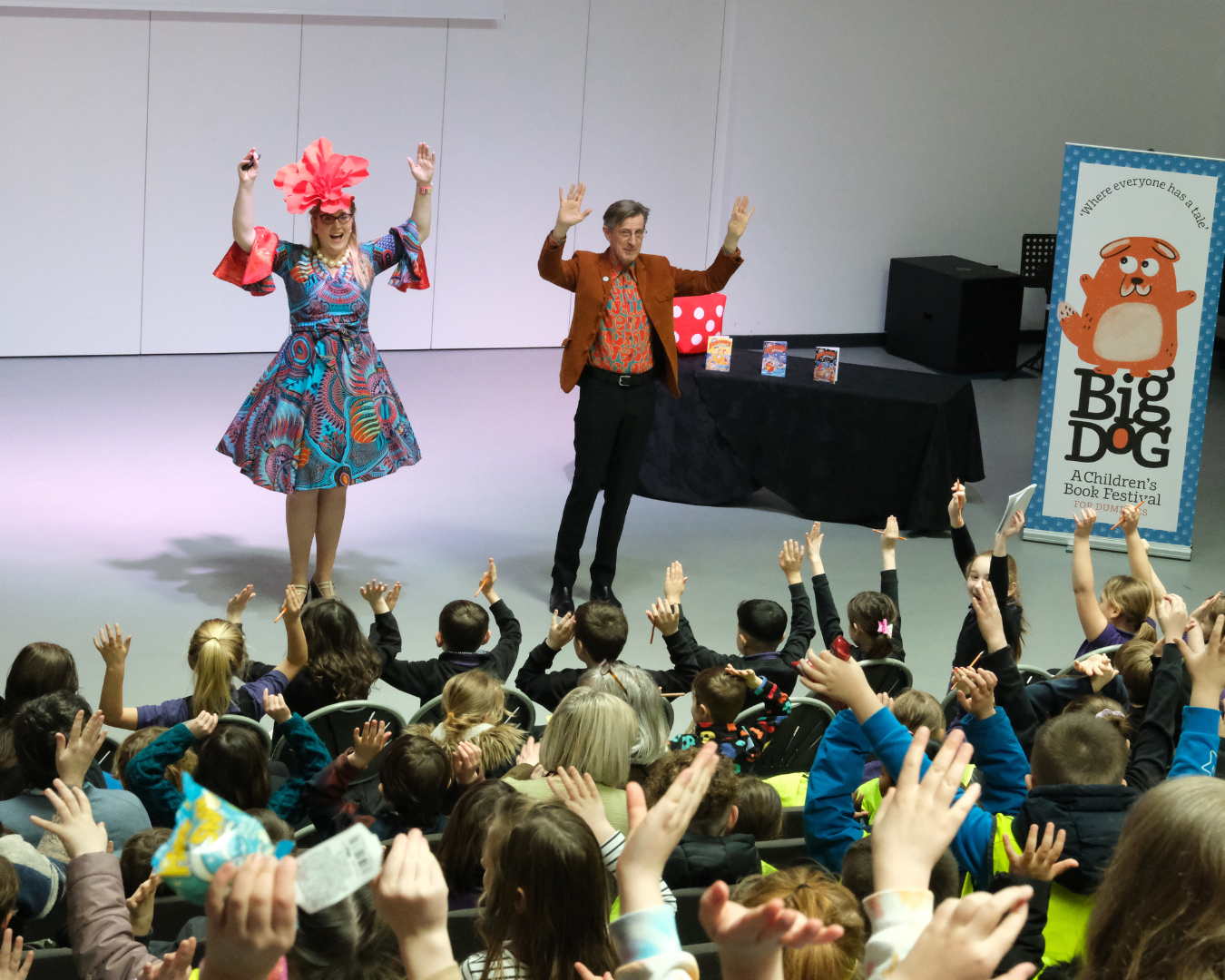 Sarah McIntyre and Philip Reeve at the Big DoG Children's Book Festival Schools Gala Day. Both are wearing flamboyant clothing, holding their arms aloft. Out of focus in the foreground is an audience of children also holding their hands up.