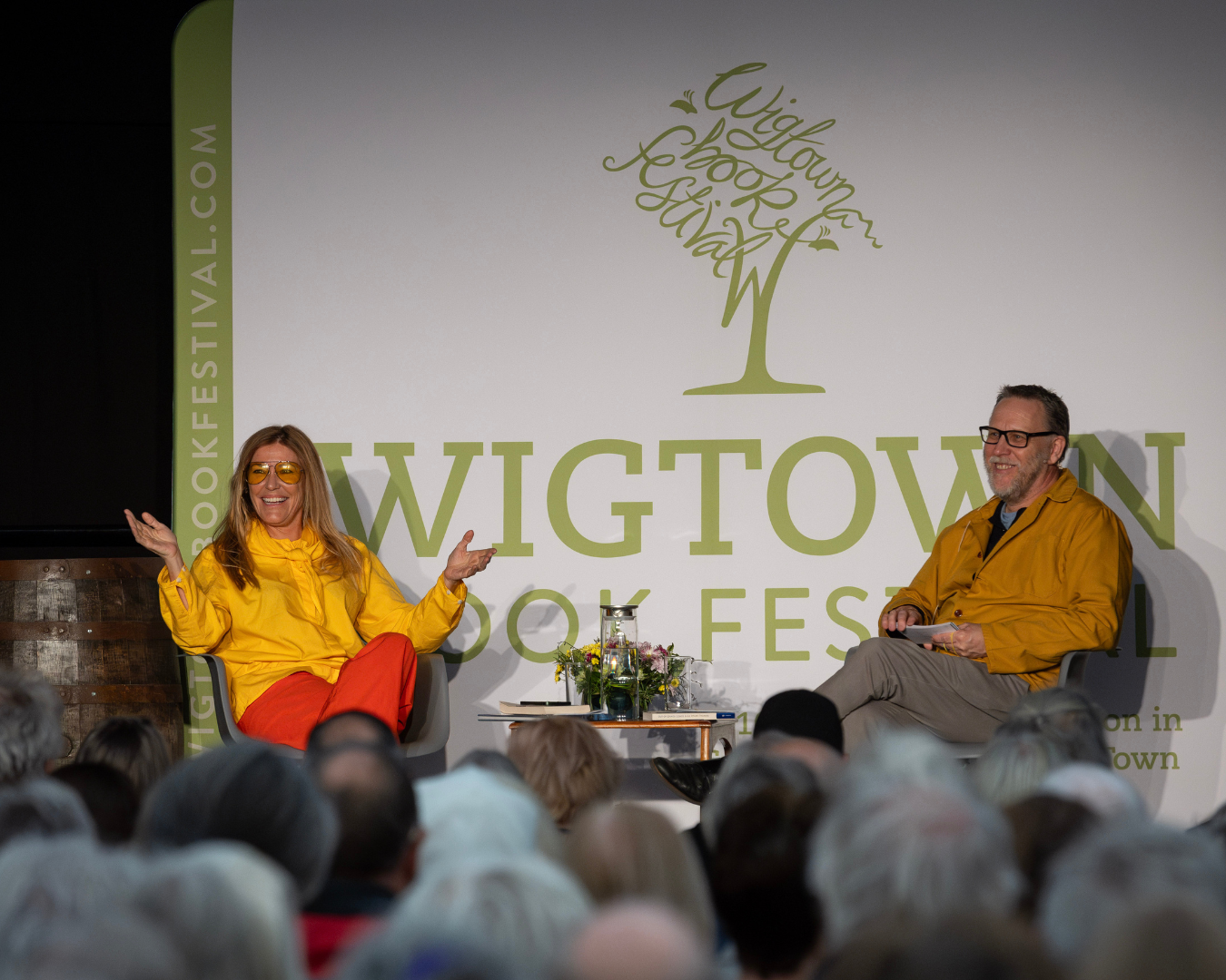 Cery Matthews, wearing a yellow jacket and yellow sunglasses, on stage at Wigtown Book Festival with chair person seated opposite, audience out-of-focus in foreground.