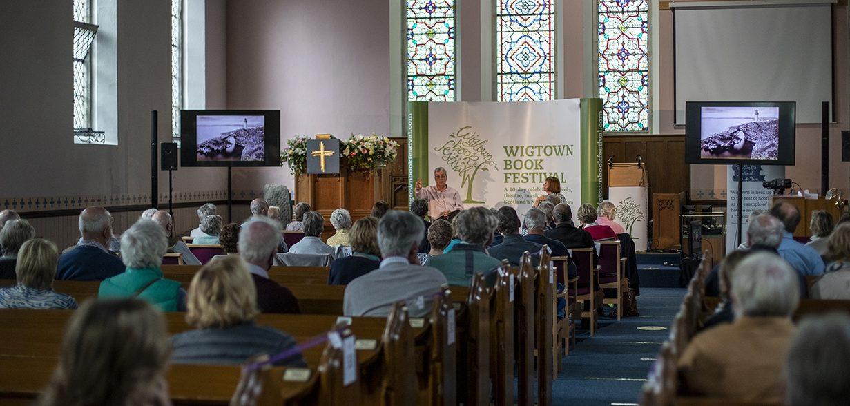 A Wigtown Book Festival event taking place in a church. The audience are sitting in the pews listening to an author talking on stage. Two screens are showing slideshows.