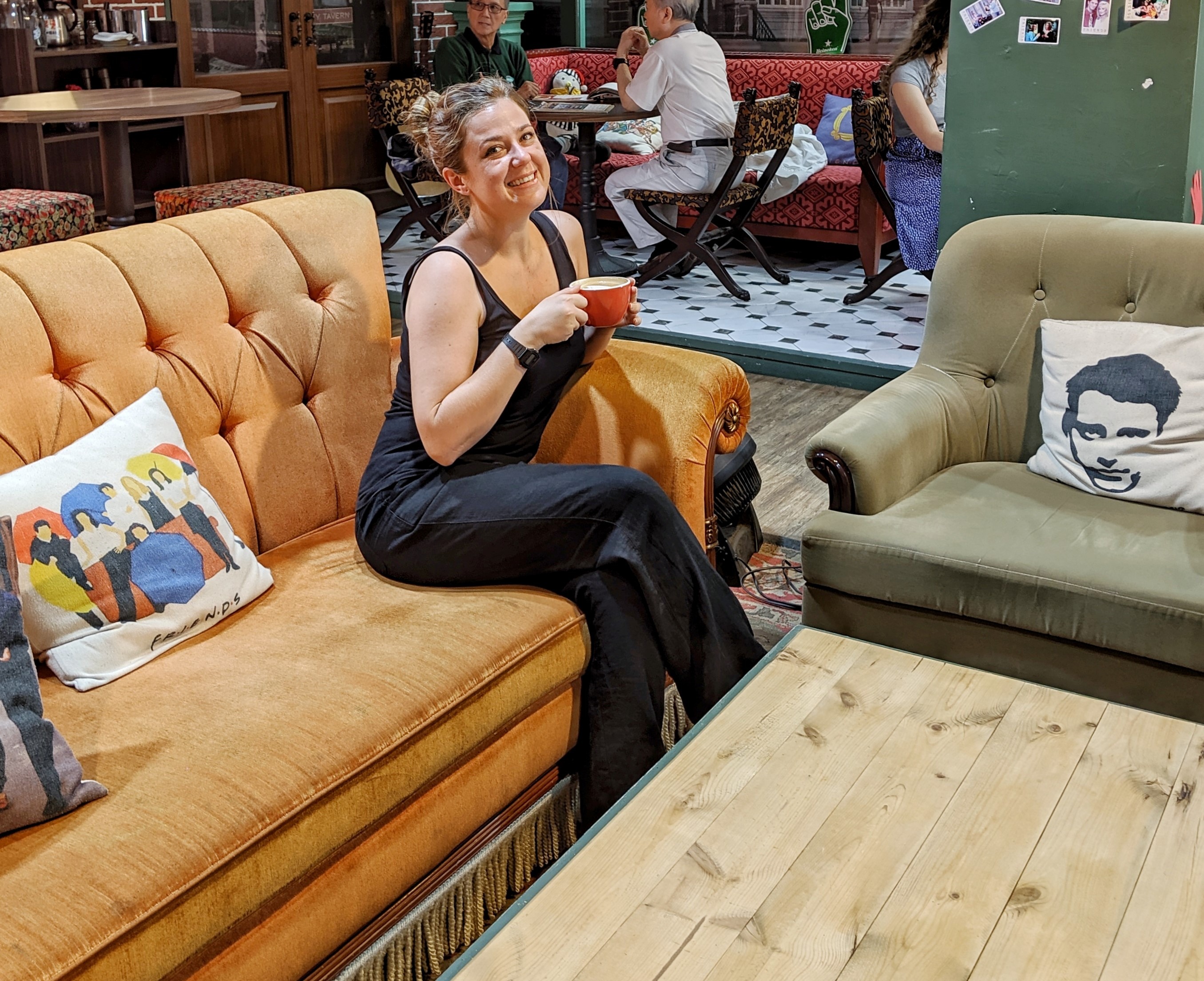 Aly Ford is sitting on an orange sofa in a coffee shop enjoying a cup of coffee. Behind her two people are talking.