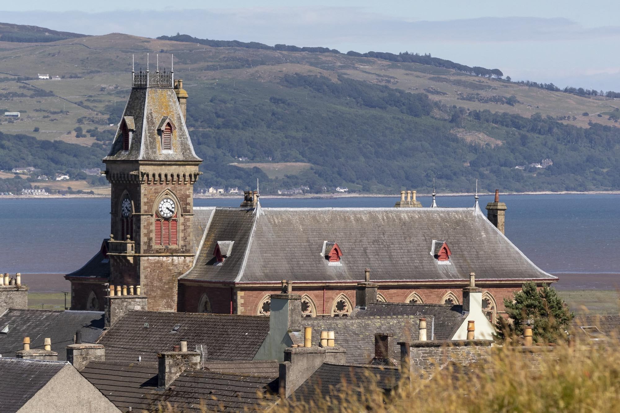 A view over rooftops of Wigtown County Buildings, the red sandstone clock tower stands prominent against a backdrop of the cree estuary and Wigtown Bay. The Galloway Hills in the distance.