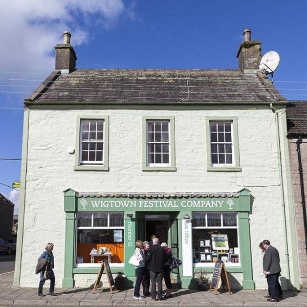 Outside the Wigtown Festival Company building on Main Street, Wigtown. It's a sunny day and a group has gathered outside the entrance of the box office, which is on the ground floor of an old building painted green.