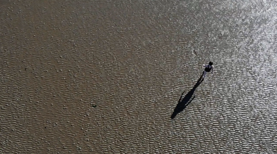 Aerial view of a lone person walking across sand in a swimming costume, the sun is casting a long shadow beside them.