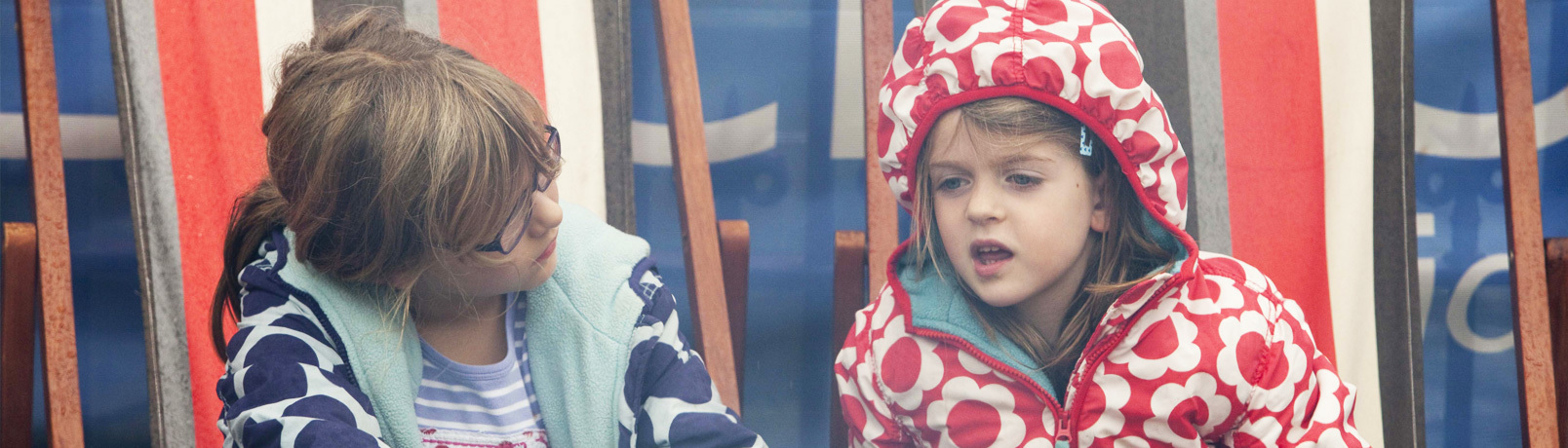 Two young children in brightly coloured coats are sitting in deck chairs talking to each other, at a Children's Book Festival event.