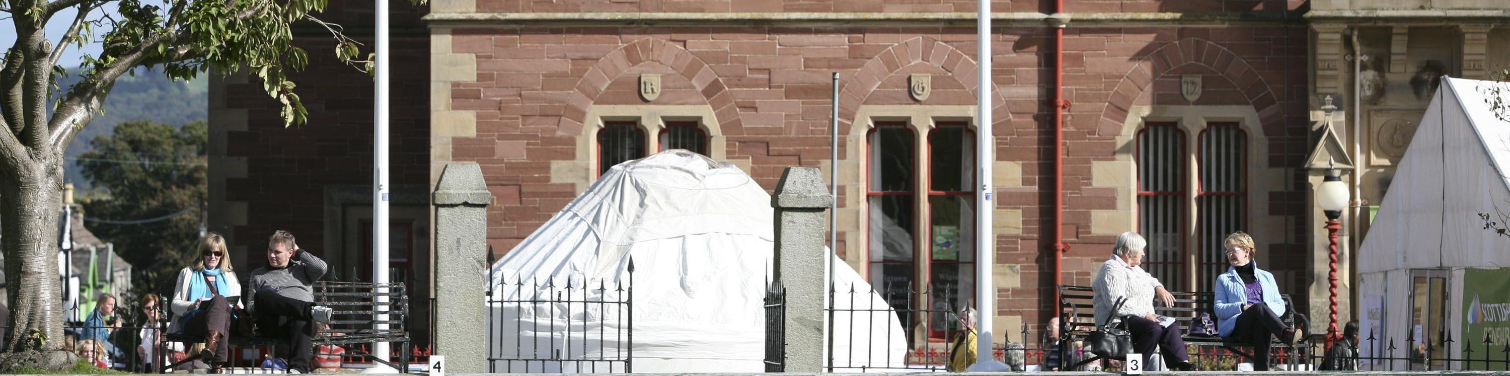 A white yurt outside Wigtown's County Buildings for a Children's Book Festival event. People are sitting on benches in the sunshine, a marquee for The Kist is situated to the right.