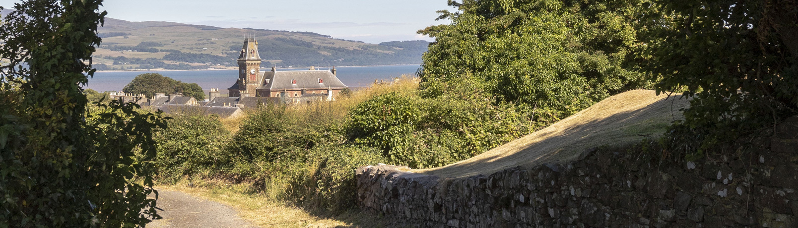 A view down a rural country lane, Wigtown County Buildings and house rooftops sit in the background against the Wigtown Bay and Galloway Hills.
