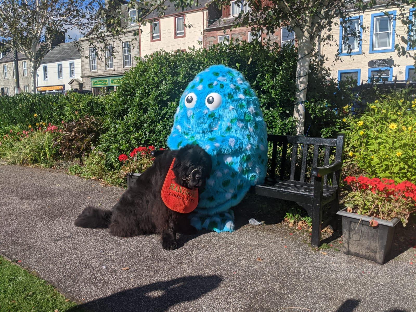 Big Wig is sitting in Wigtown Gardens with a black Newfoundland dog. Bushes, flowers and bookshops are seen behind them.