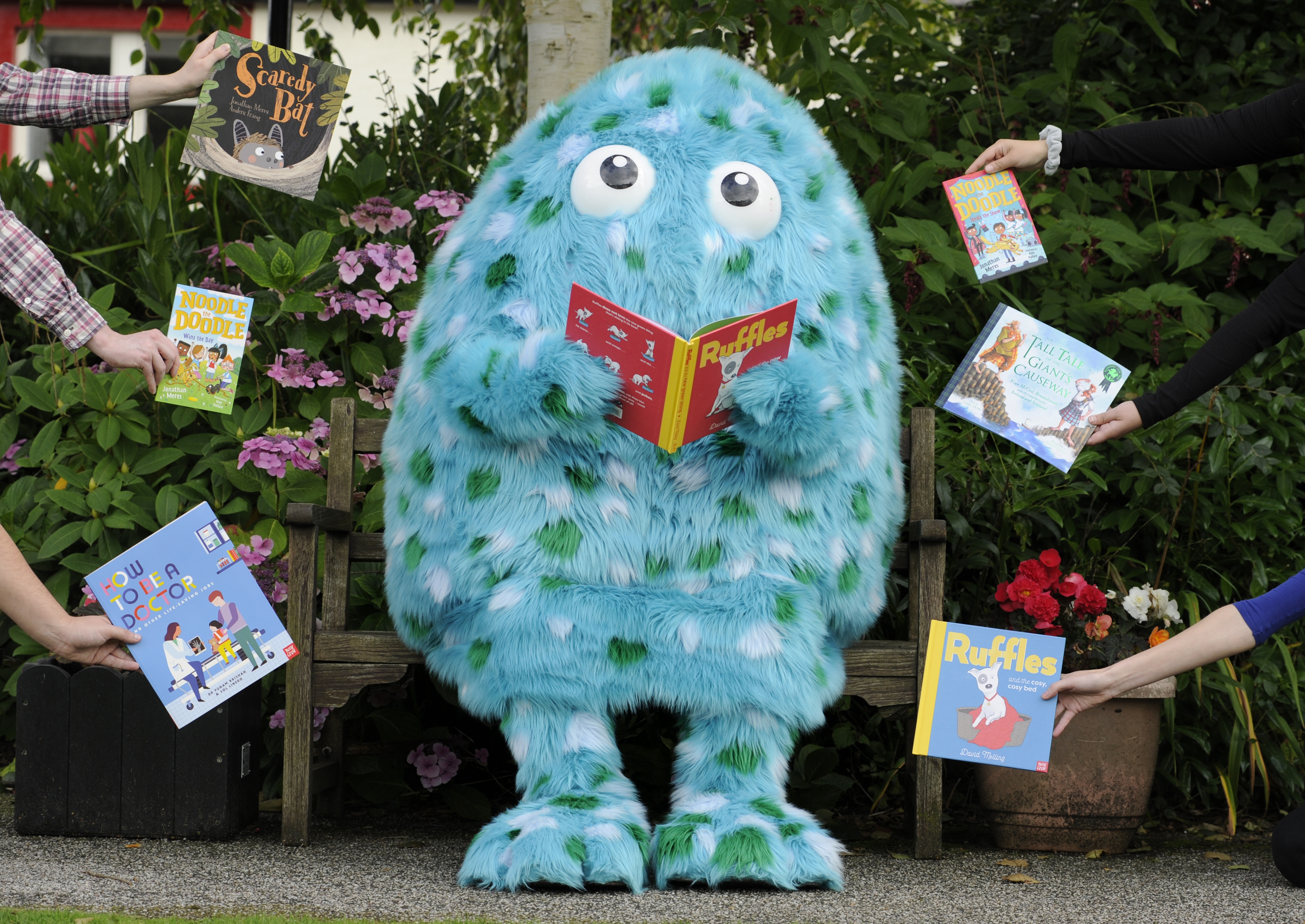 Bigwig, the Children's Book Festival mascot stands in the Wigtown Gardens holding an open book. Arms holding books are surrounding Bigwig.