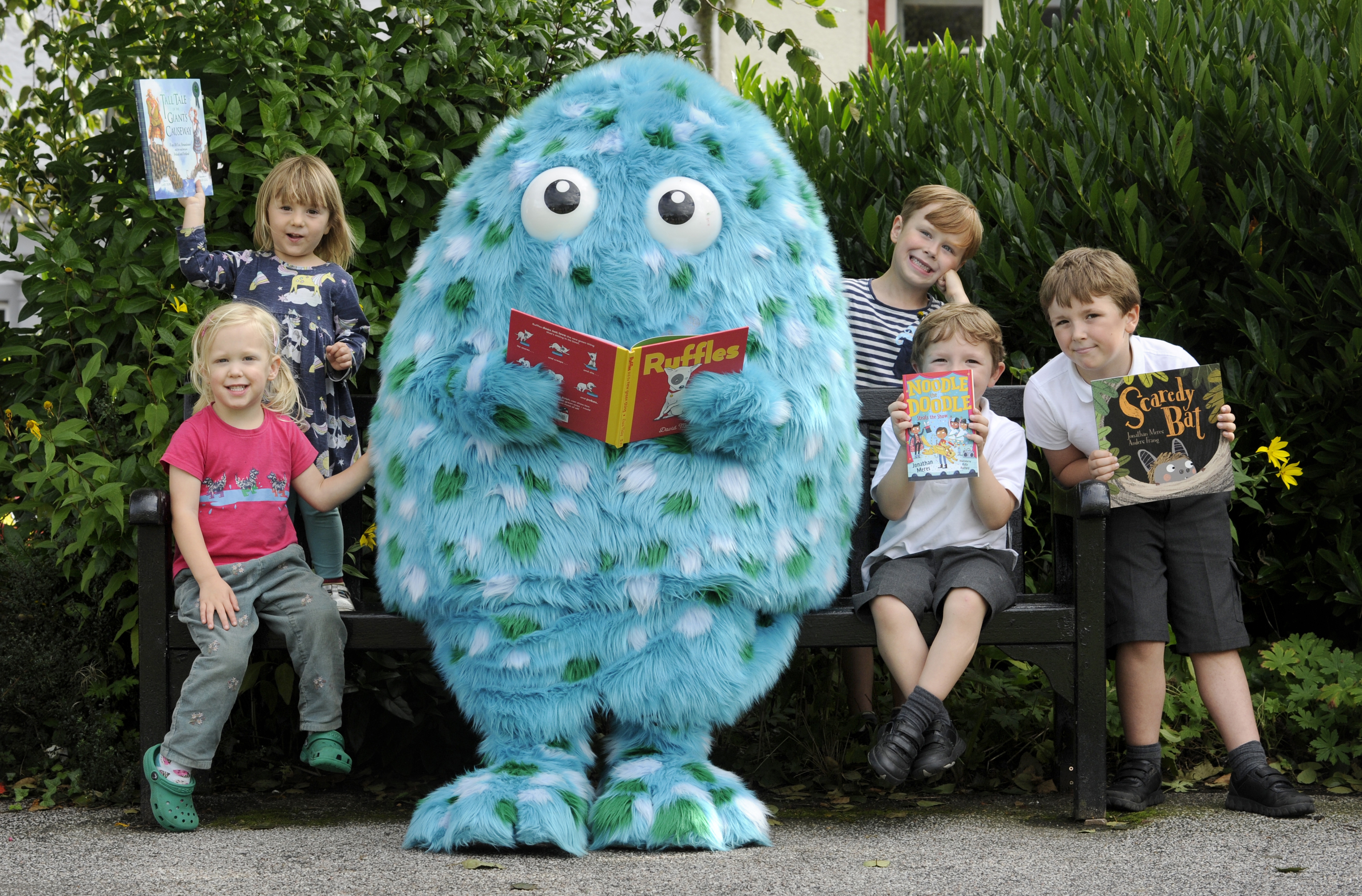 Bigwig, our children's book festival mascot, stands with various children in Wigtown gardens reading a book, The children are smiling and holding books up.