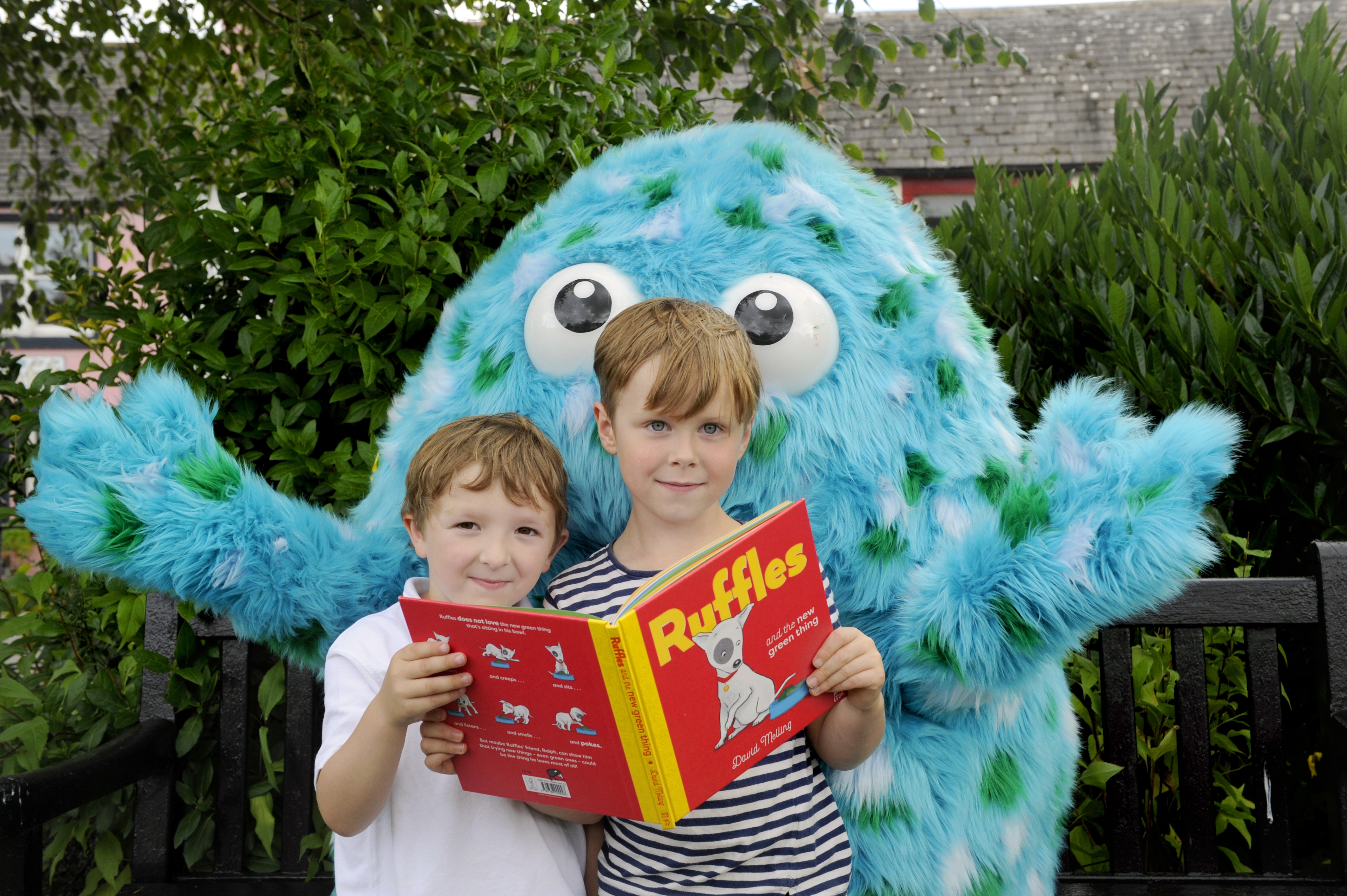 Bigwig, the Children's Book Festival mascot stands holding out his arms in Wigtown Gardens. Two young children holding a book stand in front of him.