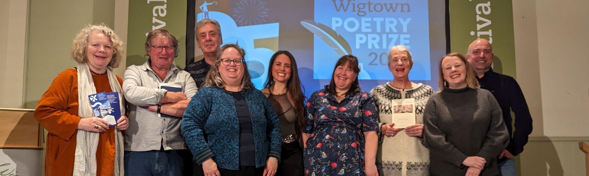 Writers and poetry 2023 winners standing on stage with Poet Hugh McMillan. A screen is behind them showing slideshows.