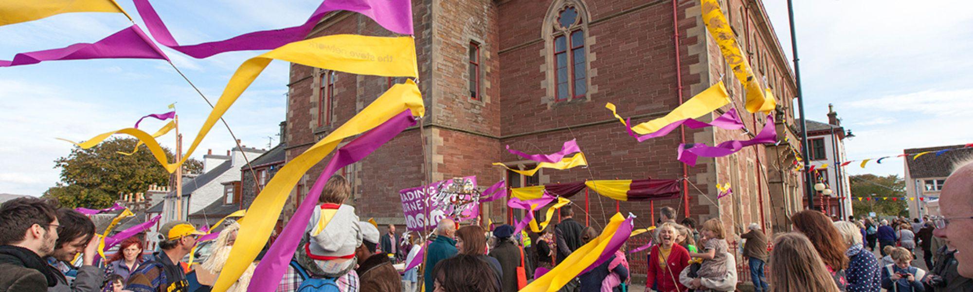 Families gathering outside the Wigtown County Buildings during a Children's Book Festival event. They are holding purple and yellow banners which are flapping in the wind.