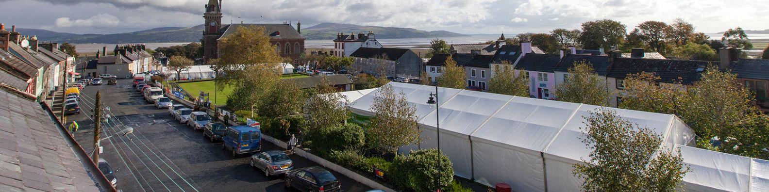 A bird's eye view of Wigtown, Scotland's National Book Town during the annual Wigtown Book Festival. The main street is lined with cars, a large marquee covers the gardens where many events are held. The County Buildings with a smaller marquee housing the Autumn Kist. Wigtown bay and the Galloway hills are in the distance.