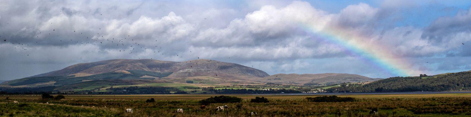 A view of the Galloway Hills over Wigtown Bay. Cumulus clouds and a partial rainbow cover the blue skies.