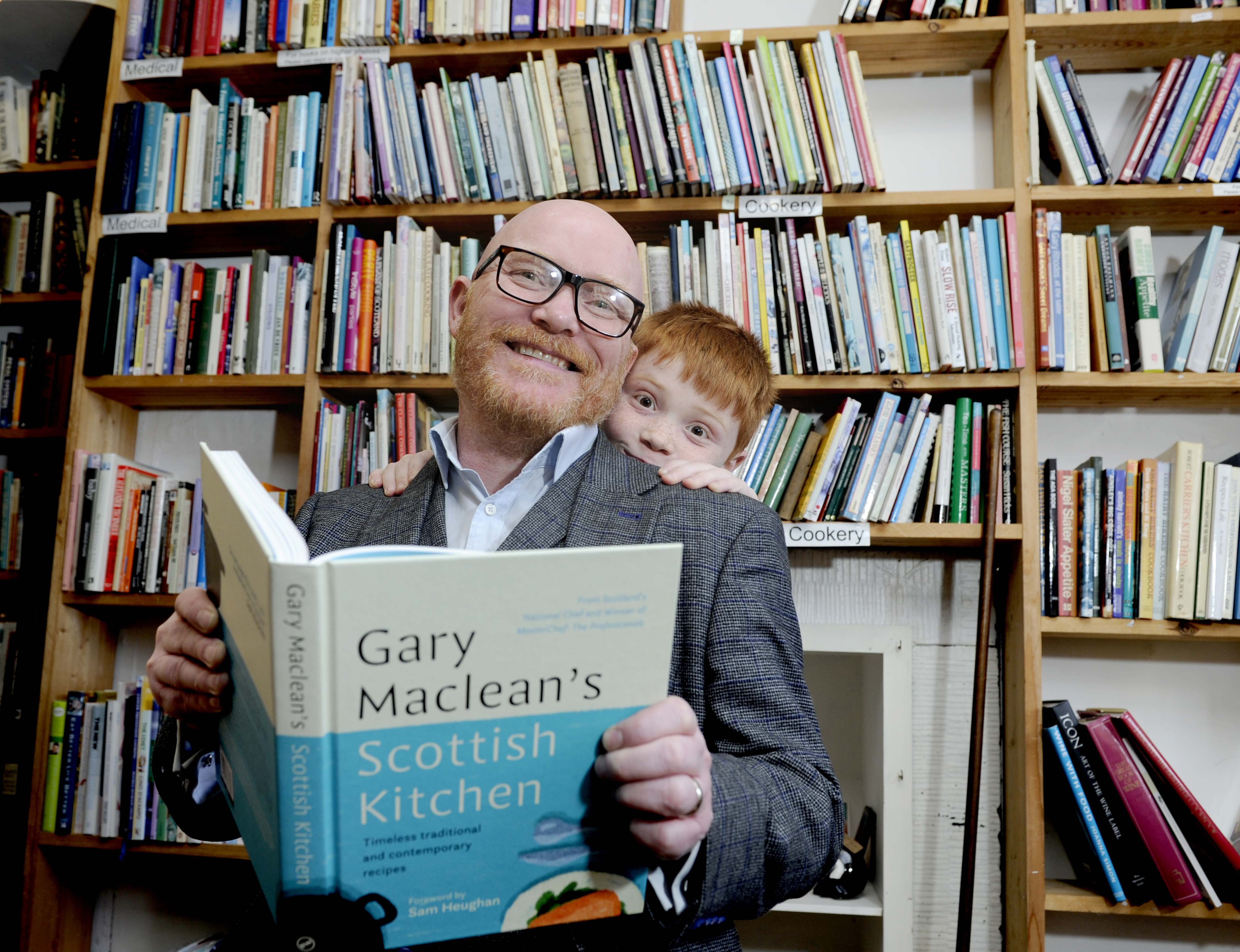 Chef Gary McClean is sitting holding his cook book in front of bookshelves full of cookery books. A young child is peering over his left shoulder.