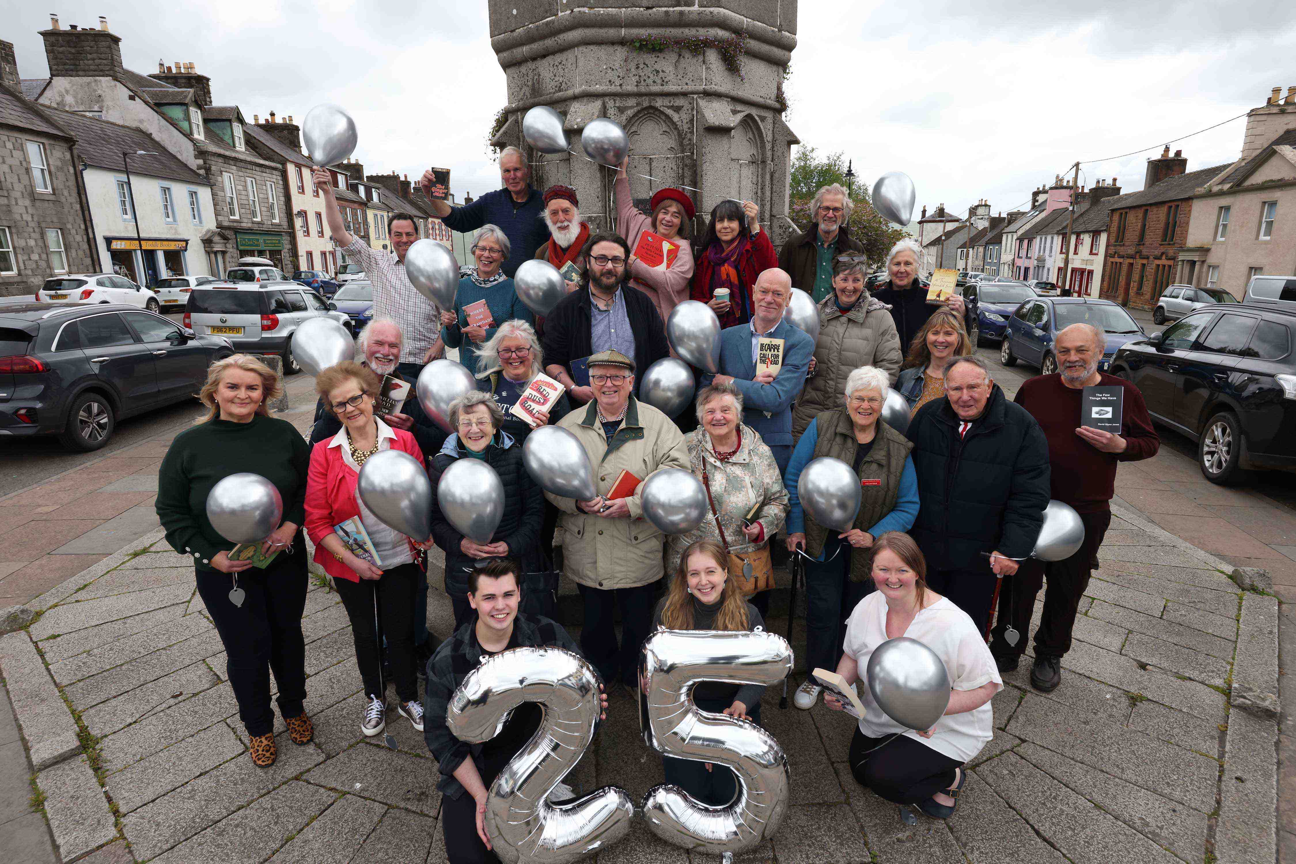 A gathering of Wigtown Festival Company staff, trustees, volunteers and Wigtown booksellers standing in front of the Mercat Cross in Wigtown, holding books and silver balloons to celebrate twenty five years as Scotland's National Book Town.