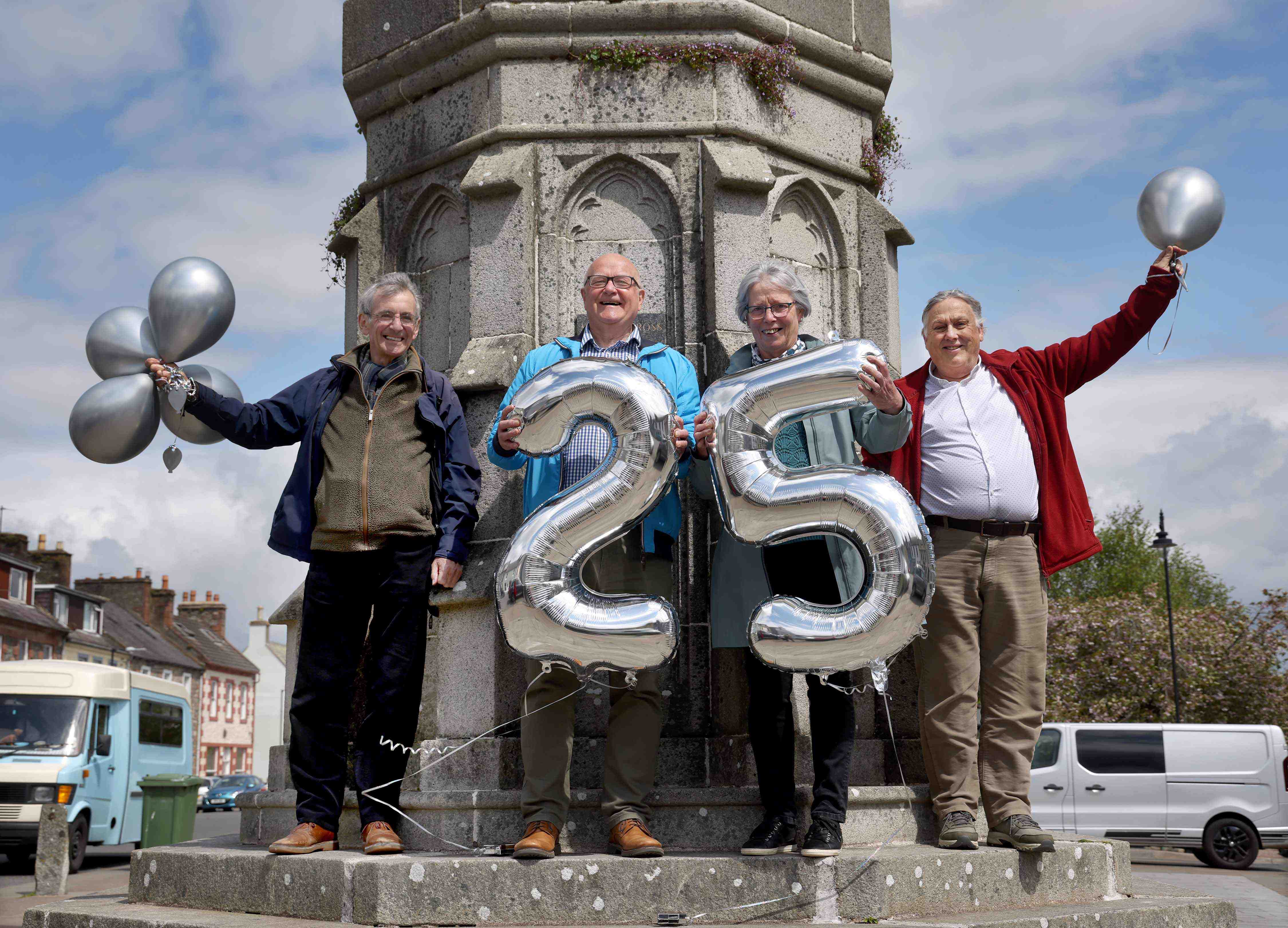 Four people standing in front of  Mercat Cross, Wigtown, holding silver balloons for the twenty fifth anniversary of Wigtown's Book Festival.