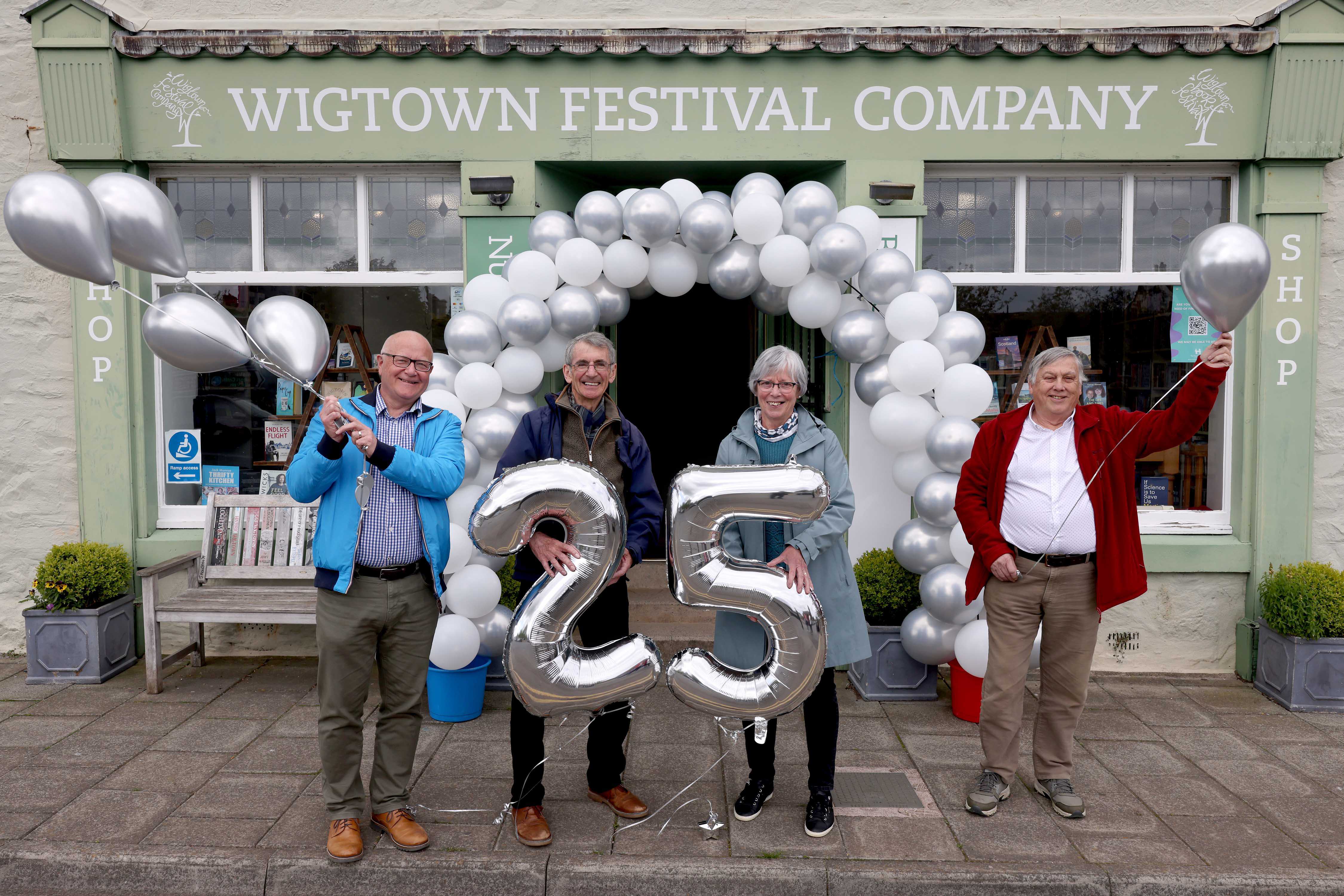 Four people standing outside Wigtown Festival Company bookshop holding silver balloons to celebrate the twenty fifth anniversary of Wigtown Book Festival.