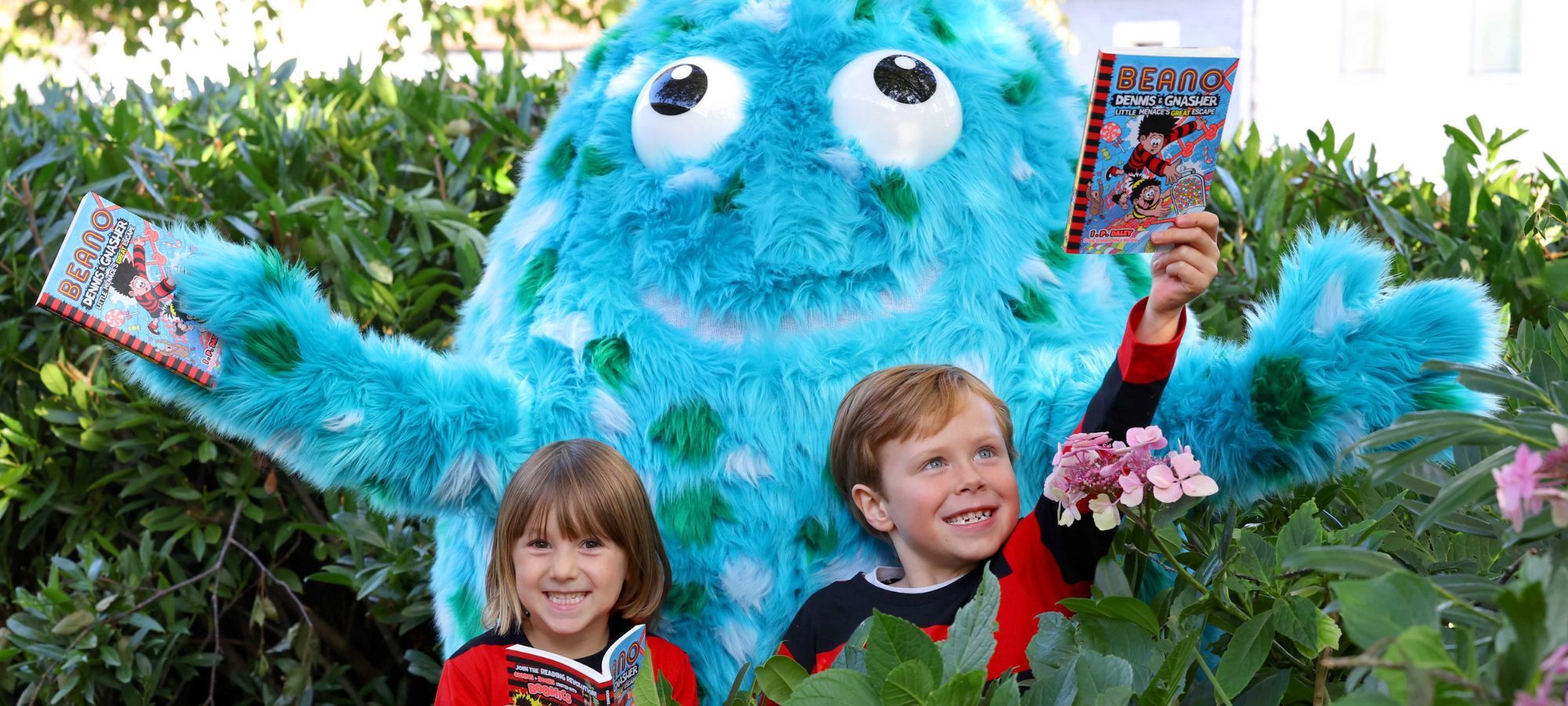 Two children in red and black striped Dennis the Menace tops hold Beano books. Big Wig, the big blue mascot, stands behind them with arms outstretched. They are in a garden surrounded by shrubbery.