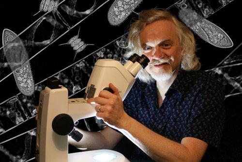 Marty Jopson sitting looking through a large microscope. Illustrations of DNA strands on the wall behind him.