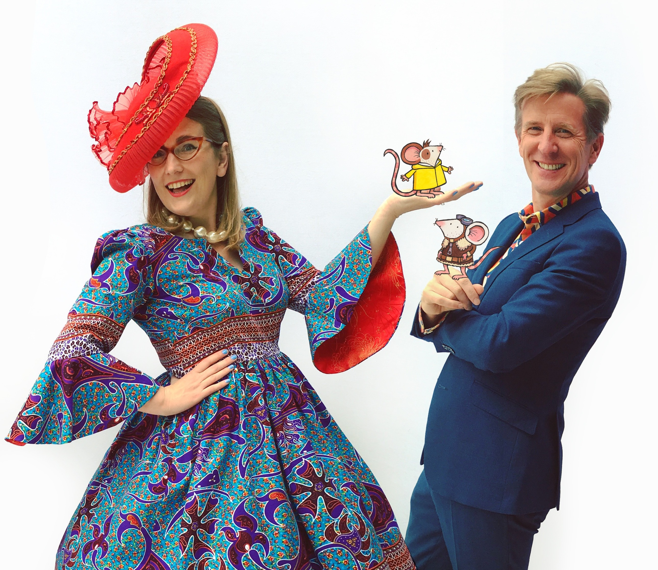 Author-illustrator Sarah McIntyre stood on the left, wearing a blue and red patterned dress with flared sleeves and a large red hat. Author-illustrator Philip Reeve stood to the right, wearing a blue suit with a red and blue patterned shirt. They each have a cartoon mouse stood on their hands, from their book Adventuremice.