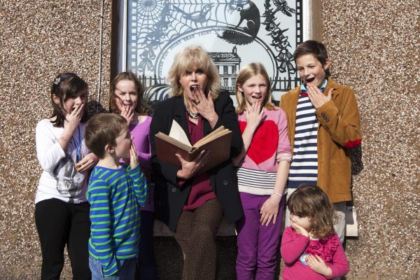 Actress Joanna Lumley standing holding an open book surrounded by six children. They all have their hands to their mouths and are gasping. Behind them a pebble dashed wall and a window with a black and white etched picture featuring a house, a fox, a bird, flowers and a large cobweb in the left hand top corner.