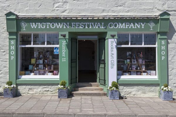 Wigtown Festival Company, shop and box office. Cream painted building with green trim. Four planters with flowers stand outside.