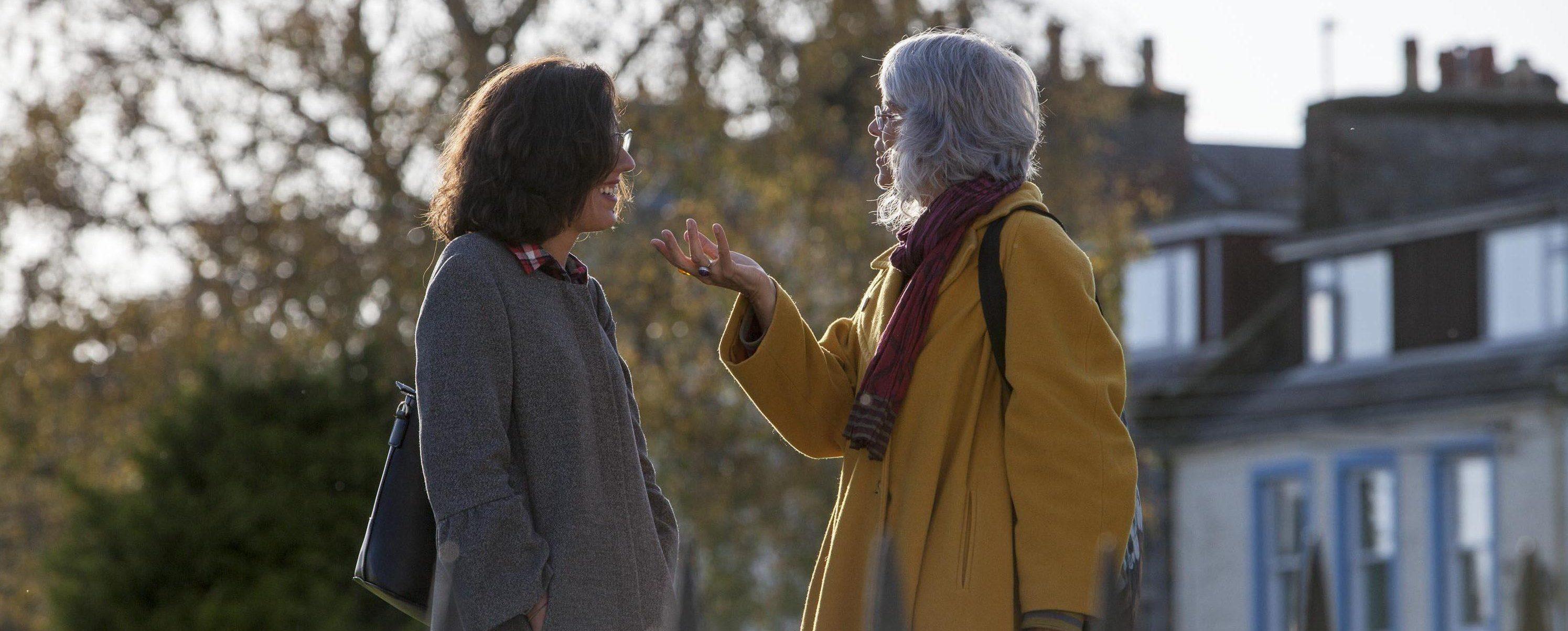 Two people standing facing each other talking, houses and trees behind them.