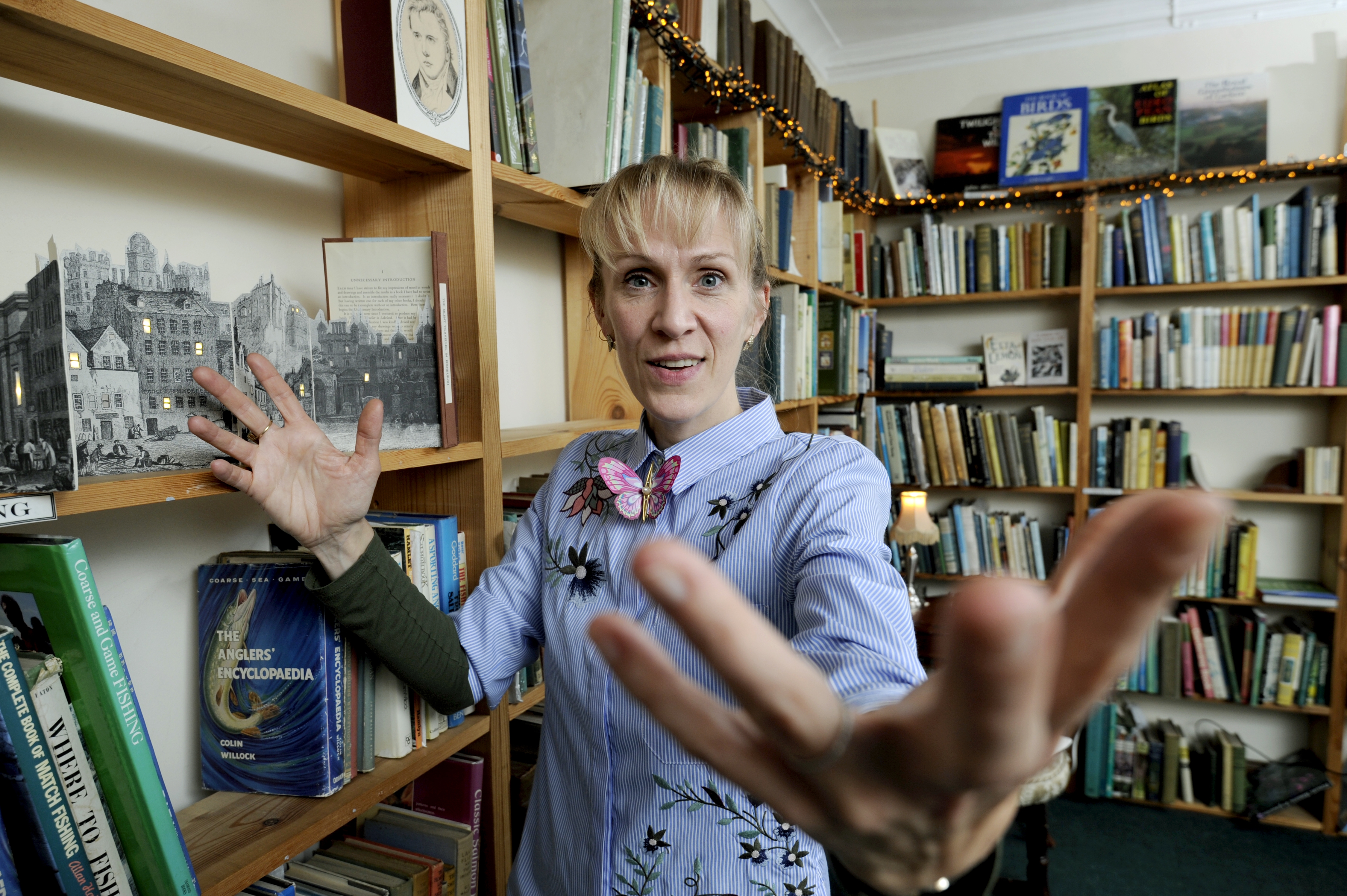 A person is standing in a bookshop gesturing with their hands. The bookshelves are full of books and fairy lights adorn the top shelf.