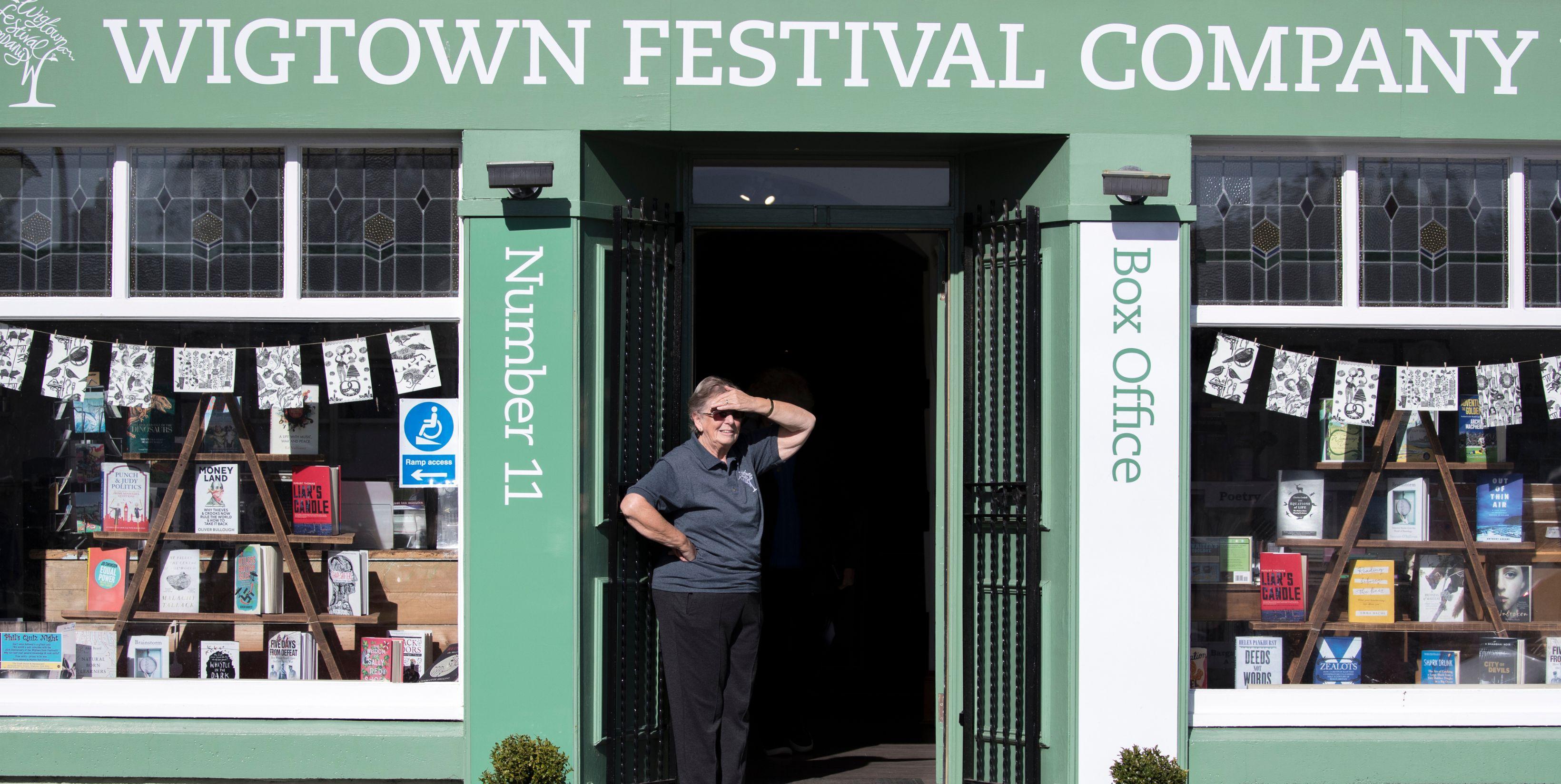 Wigtown Festival Company bookshop and box office. The windows are dressed with books and bunting. A volunteer looks out of the door with their hand shielding their eyes from the sunshine.
