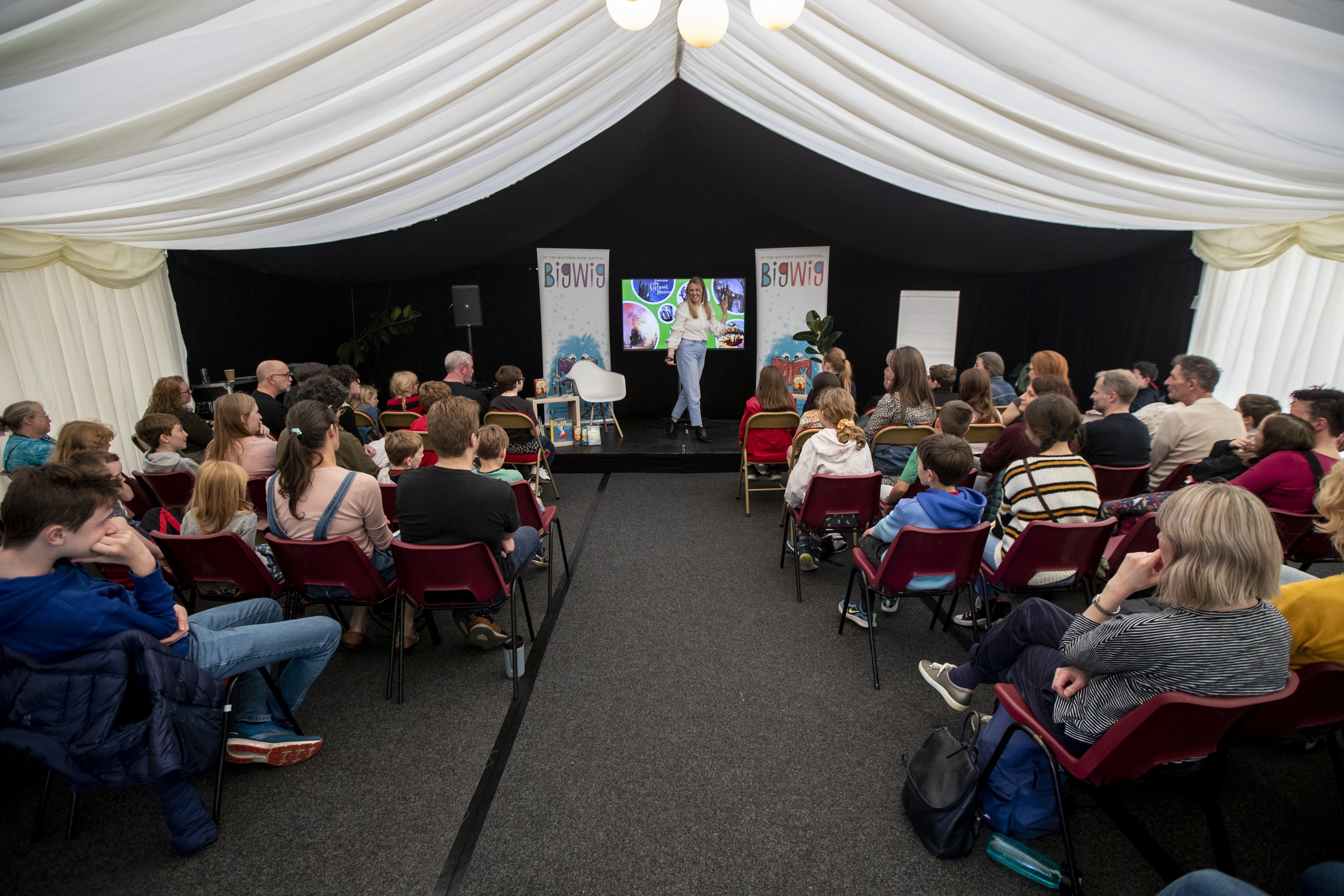 Children's author Vashti Hardy animatedly talking on stage at her event at Wigtown Book Festival. The audience sit in front of her listening. Above her white material drapes the ceiling with a chandelier hanging down.