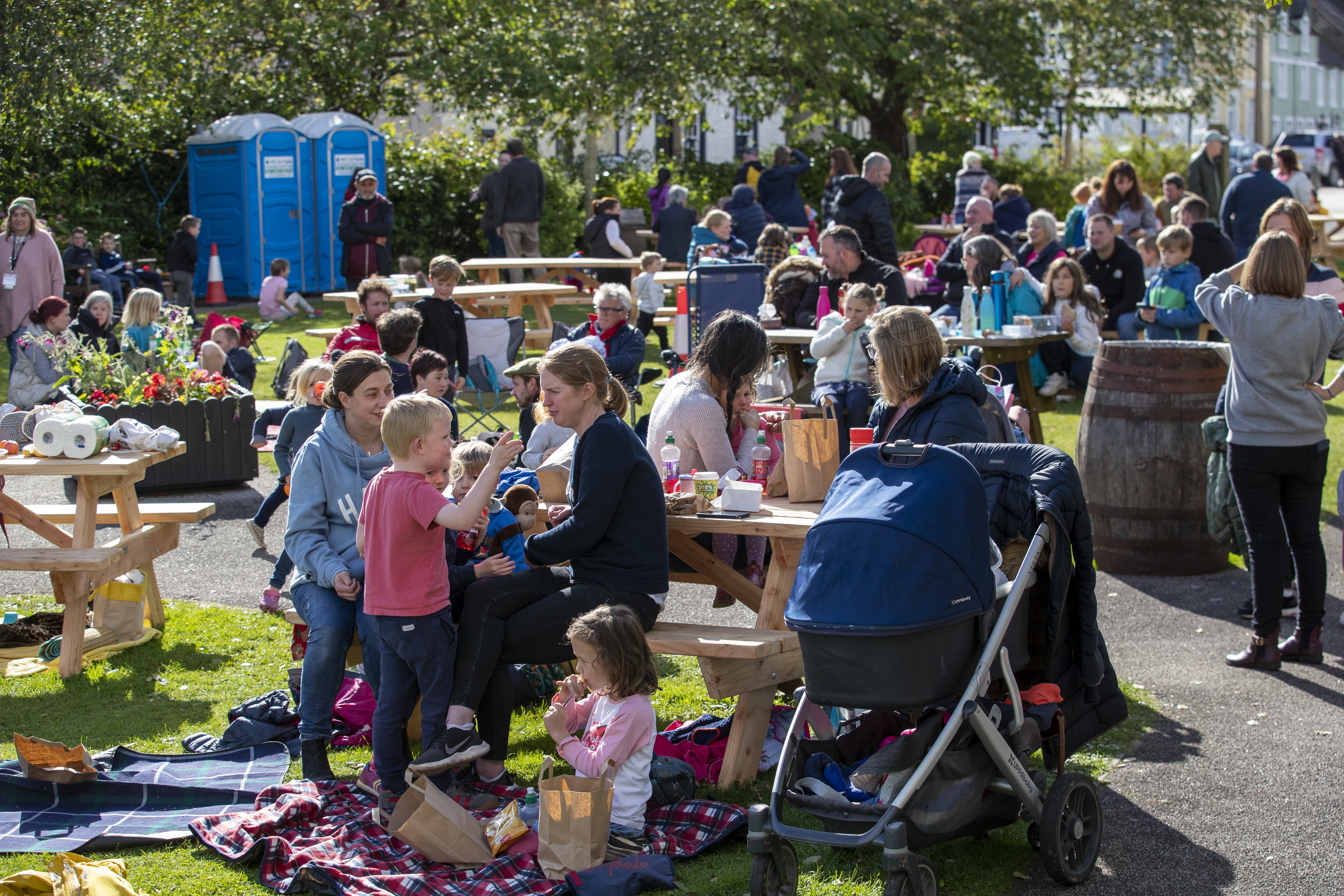 Families sitting eating and drinking at picnic benches in Wigtown Gardens during the Wigtown Book Festival. The sun is shining.