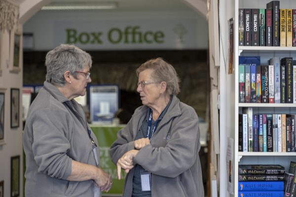 Two Wigtown Book Festival volunteers talking to each other in the box office. One is pulling back their sleeve to see their watch.