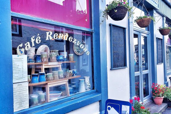 Side view of Cafe Rendezvous in Wigtown. White building with blue window and door trim. Hanging baskets over the door, red geraniums  in pots outside. The window display features pottery items for sale.