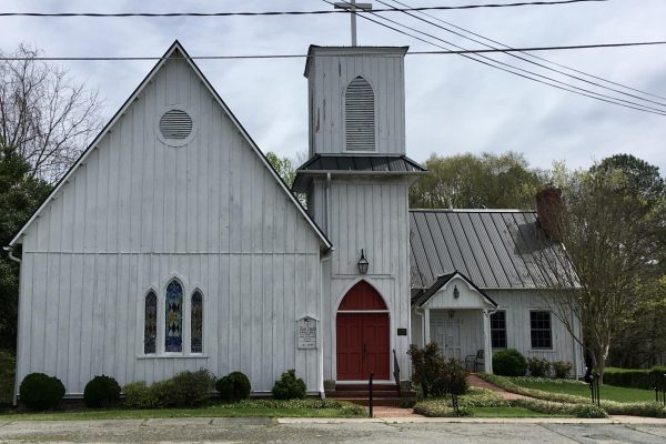 A white wooden framed church sits on the road in North Carolina. The church has a red door and a cross above the tower.