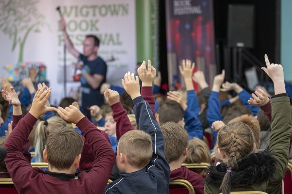 School children are seated in the main marquee raising their hands to answer questions from an author on  stage, at a Children's Book Festival event.