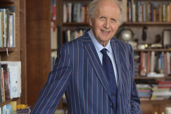 Alexander McCall Smith is standing in a room surrounded by bookshelves full of books.