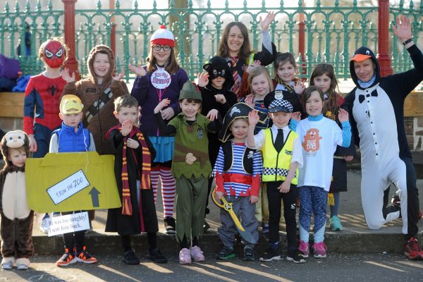 A group of children dressed up as various book characters stand waving and smiling at a Children's Book Festival event. Green railings are behind them.