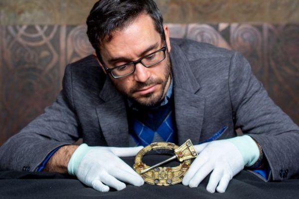 Adrián Maldonado is wearing white gloves holding a brooch from a collection at the National Museum of Scotland.