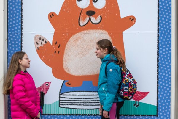 Two children stand talking outside Big DoG HQ in front of a large window decoupage featuring the character Big DoG, an orange dog, with the caption 'Where everybody has a tale' above.