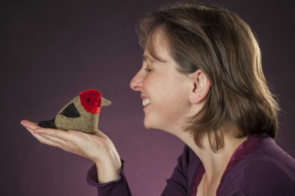 A side view of storyteller and puppeteer Ailie Finlay holding a bird in her hand up towards her mouth and smiling.