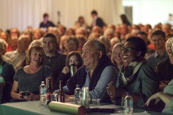 A gathering of people enjoying an event at Wigtown's Book Festival.  In the fore front three people are sitting at a table with drinks and papers laughing.