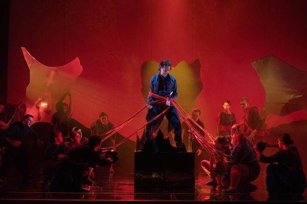 Daniel Keating Roberts as Orfeo with Scottish Opera Young Company in Orfeo Euridice Scottish Opera 2019 . The principal is standing on a plinth  bound by ropes held by various cast members. The stage is lit by red lights to infer flames.