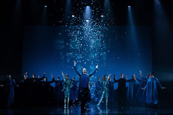 Daniel Keating Roberts standing on stage with his hands in the air, as Orfeo with Scottish Opera Young Company in Orfeo Euridice Scottish Opera 2019. The stage is lit in blue and white confetti is falling down on the principal.