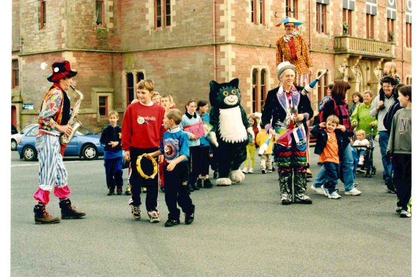 Children and families are walking in front of Wigtown County Buildings amongst entertainers wearing colourful clothes, playing instruments and walking on stilts during a Children's Book Festival event.