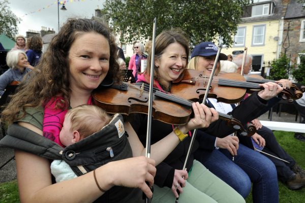 Beth Please and others sit playing their fiddles in Wigtown Gardens at an outdoor event during Wigtown Book Festival. Beth has a baby in a front sling. Other musicians in the background standing talking amongst themselves.