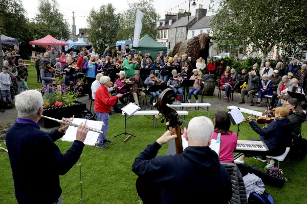 The Fiddlers Fair, an outdoor event in Wigtown gardens as part of the Wigtown Book Festival. The fiddlers sit in a semi-circle, in front of them is a flute player, the double bass, keyboard and accordions. The conductor stands in the middle with her baton raised. A mixed audience sit behind and to the right listening to them play.