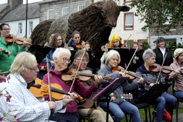 The Fiddlers Fair at Wigtown Book Festival. Various people are sitting playing their violins in the gardens.  Kelton the bull, larger than life wicker sculpture is placed behind them.