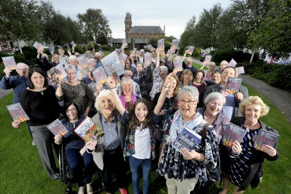 Volunteers and staff from Wigtown Book Festival stand together in the gardens holding the program brochure. Wigtown County Buildings in the background.